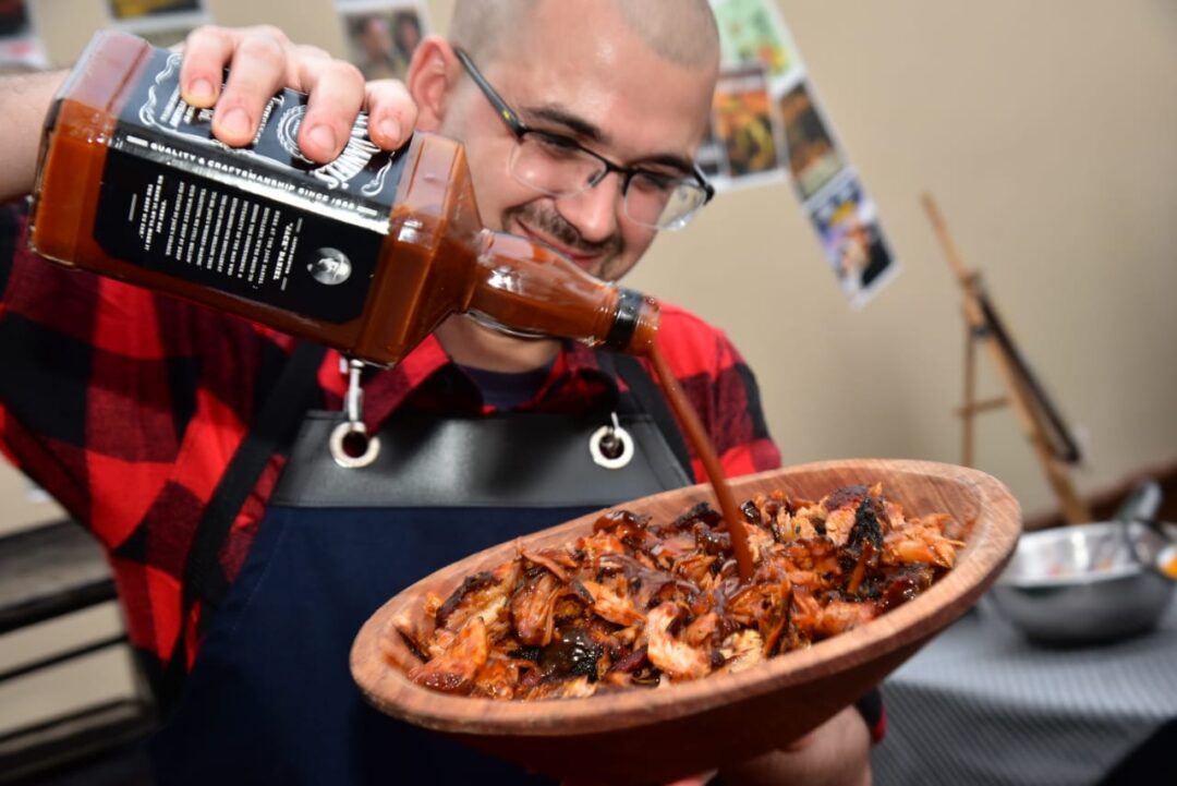 Homem de óculos e camisa xadrez vermelha despeja molho barbecue sobre carne desfiada em tigela de madeira