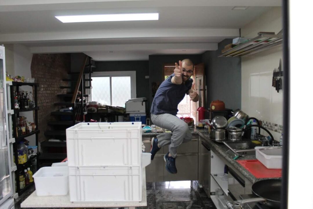 Homem de camisa azul e calça cinza salta em cozinha, fazendo sinal de positivo entre caixas plásticas e panelas