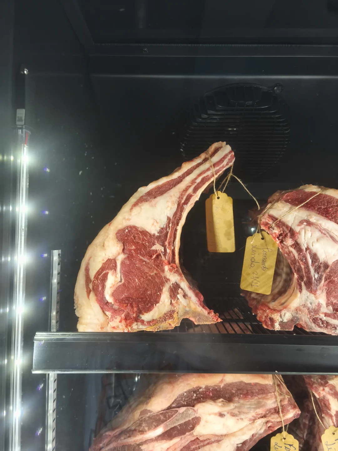Peças de lombo bovino com osso penduradas em câmara refrigerada com etiquetas amarelas escritas à mão