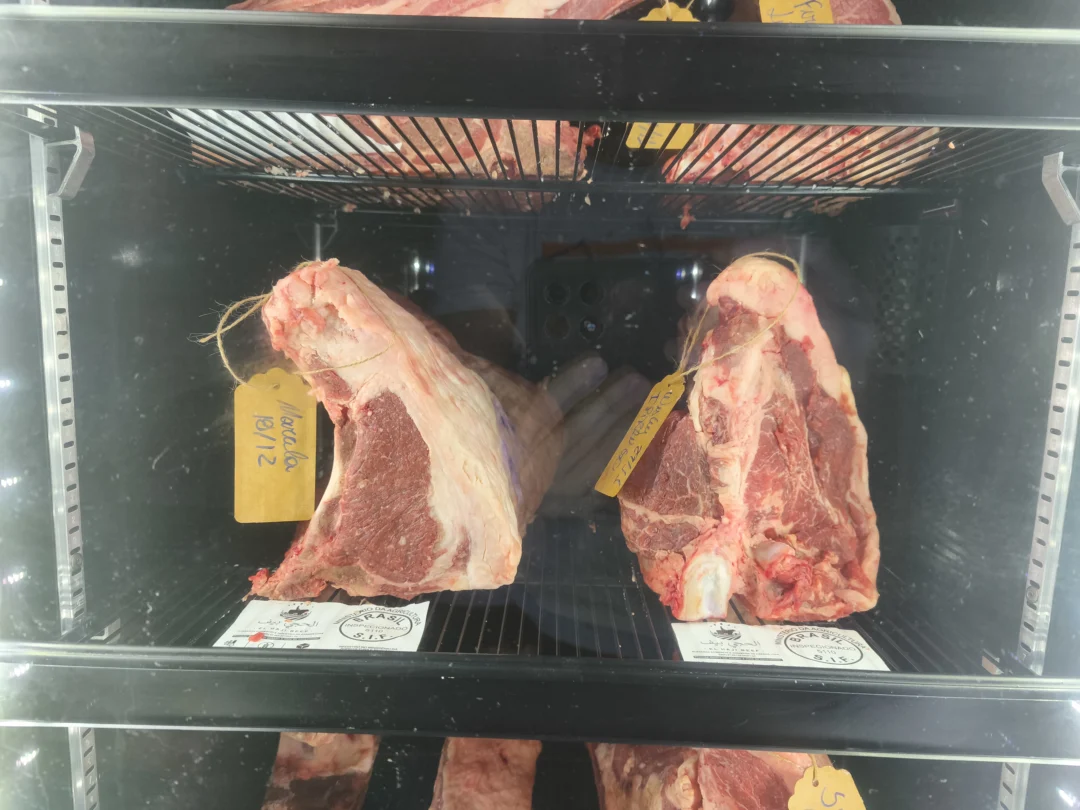 Duas peças grandes de carne bovina com osso penduradas em câmara refrigerada com etiquetas amarelas