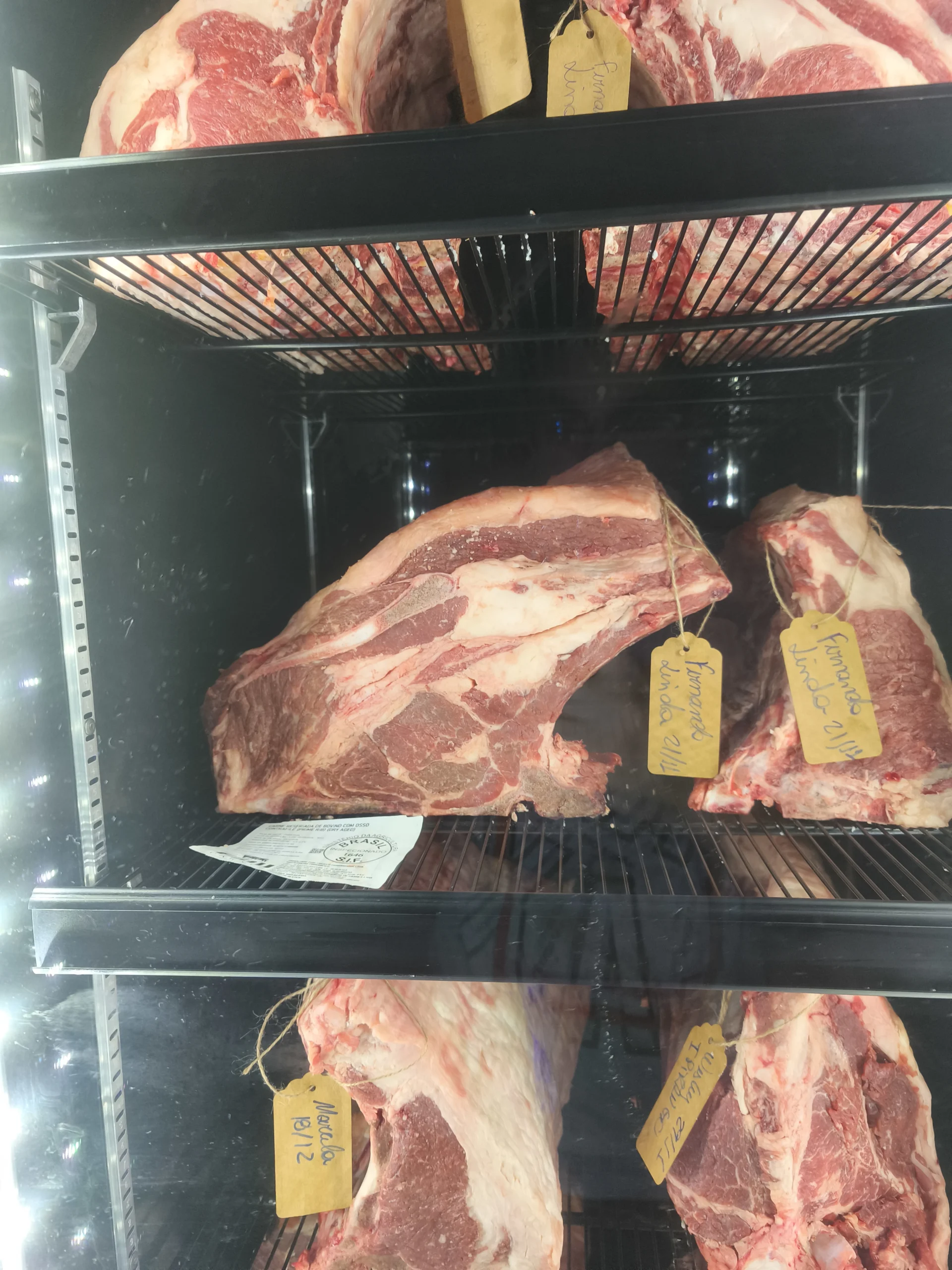 Peças de carne bovina maturando em câmara de vidro, com etiquetas amarelas escritas à mão